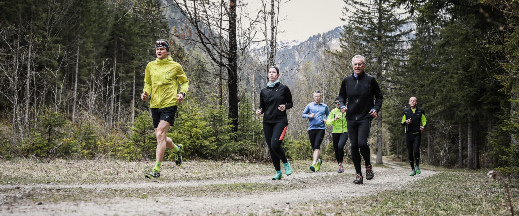 Gruppe von Jogger:innen läuft auf Waldweg durch Berglandschaft, umgeben von Bäumen und Natur.