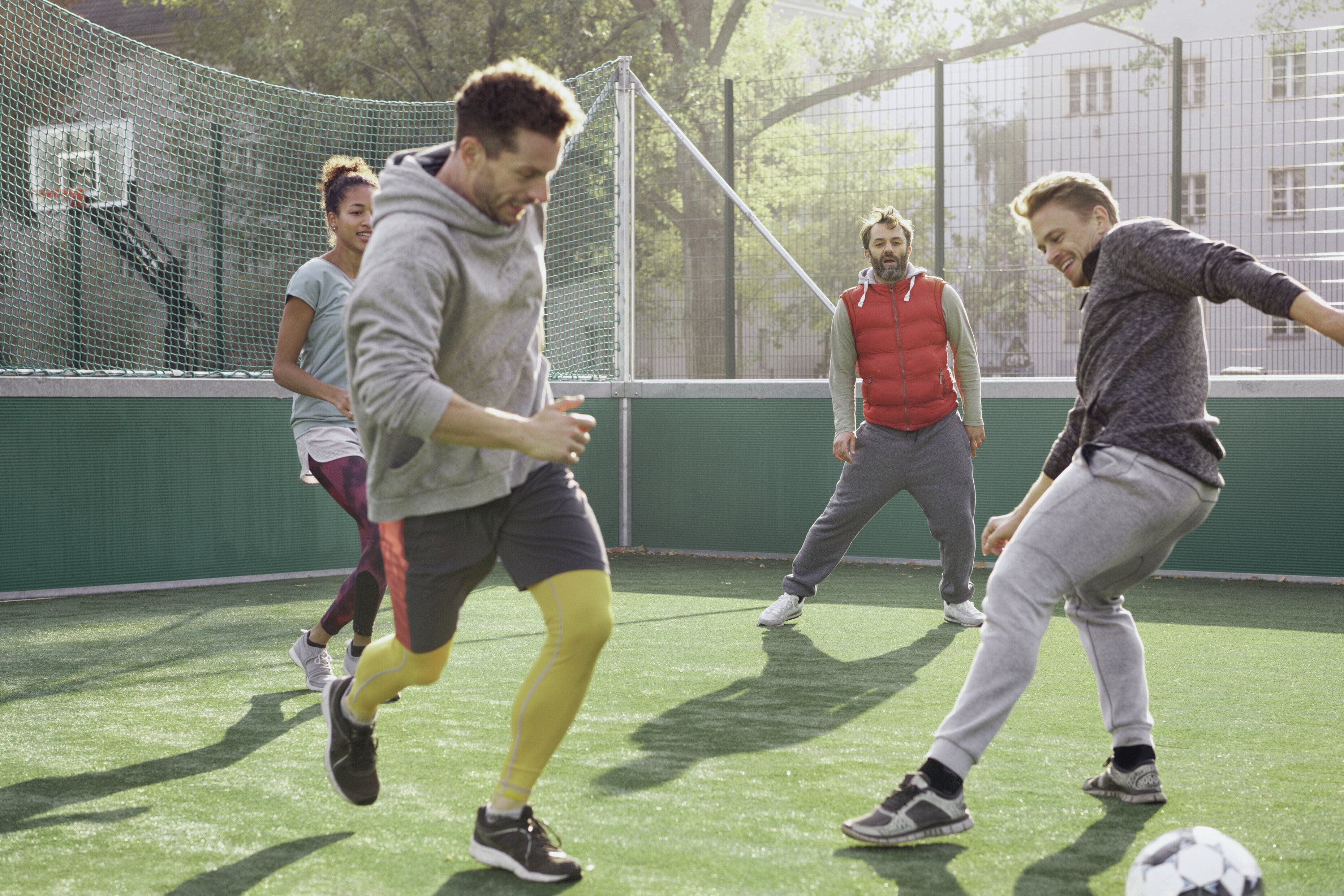 Vier Erwachsene spielen lachend Fußball auf einem kleinen Kunstrasenplatz bei Sonnenschein.