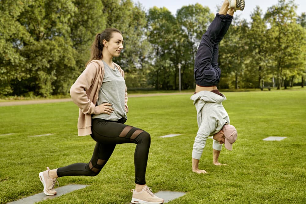 Frau macht Ausfallschritt, Mann Handstand - beide trainieren sportlich im Park bei grünem Wetter.